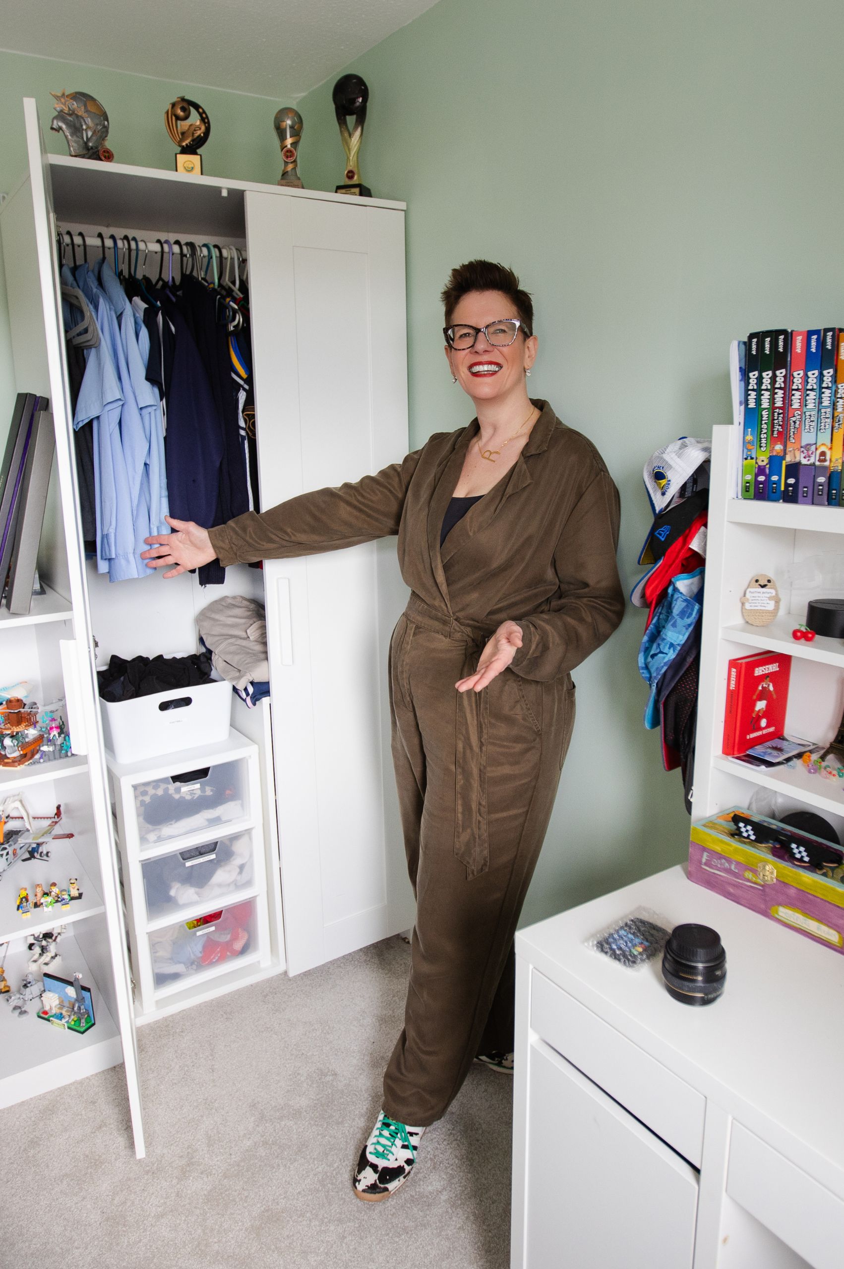 Roxy Decluttering Bedroom Roxy of Declutter & Reorganise with Roxify smiling and gesturing proudly toward a neatly organized open white wardrobe in a child's bedroom. She is wearing a brown jumpsuit and glasses, standing on a grey carpet surrounded by tidy white shelving units filled with arranged clothes, books, caps, and toys.