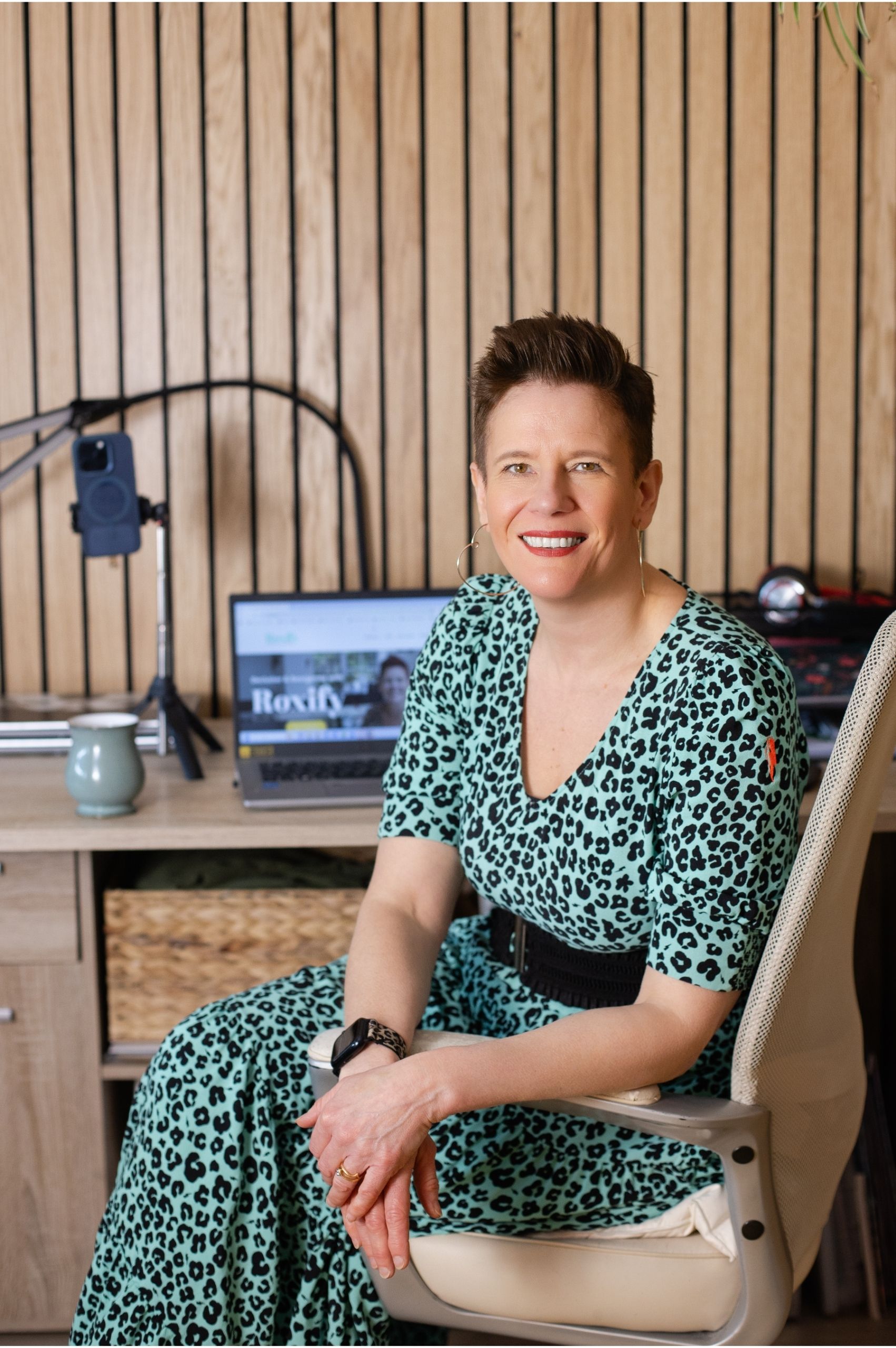 Roxy of Declutter & Reorganise with Roxify smiling as she sits in a cream office chair at her workspace. She is wearing a turquoise leopard print dress with a black belt. Behind her is a wood-slatted wall and a desk holding a laptop displaying her "Roxify" website, a mug, and a smartphone mounted on a tripod